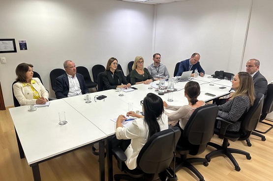 Foto tirada de pessoas sentadas em cadeiras estofadas pretas ao redor de uma mesa branca. Quem está sentado ao lado esquerdo da mesa está de frente para a fotografia.