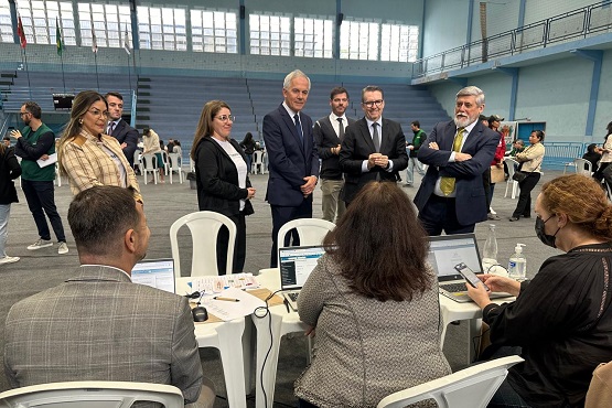 Em um ginásio, três assistentes estão sentados de costas para a foto, cada um em uma  cadeira de plástico e uma mesa na frente, com um computador sobre ela. Ao fundo estão 4 homens engravatados e duas mulheres em pé.