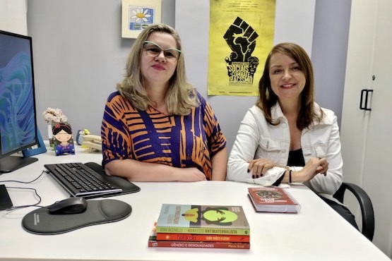 Duas mulheres estão em uma sala sentadas cada uma sobre uma cadeira e em frente a uma mesa.