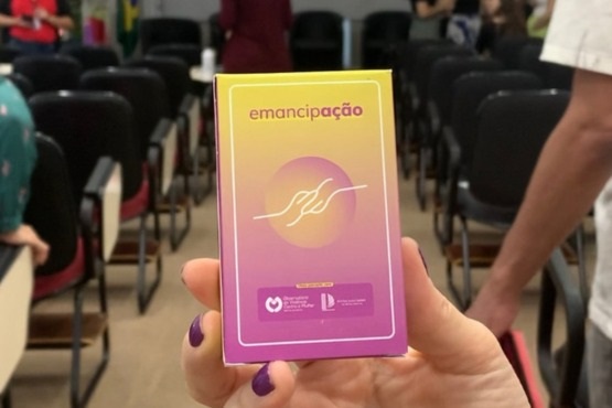 Mão com unhas roxas segura uma caixa retangular na coloração de um degradê amarelo e roxo claro. No topo está escrito “emancipação” em letras roxas. Ao centro está um desenho gráfico em branco envolto por um círculo das cores do degradê. No final está os logos dos apoiadores do projeto.