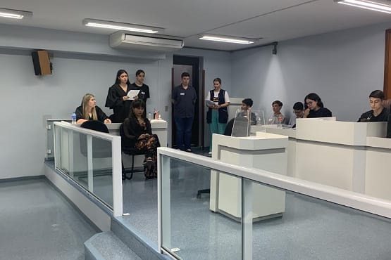 Alunos dentro do Tribunal do Júri de São Bento do Sul. Ao lado esquerdo da imagem, uma aluna de pé lê uma folha de papel enquanto duas mulheres ao seu redor estão sentadas. Ao fundo, mais três pessoas estão de pé. No lado direito, cinco alunos estão sentados em mesas brancas destinadas aos juízes.