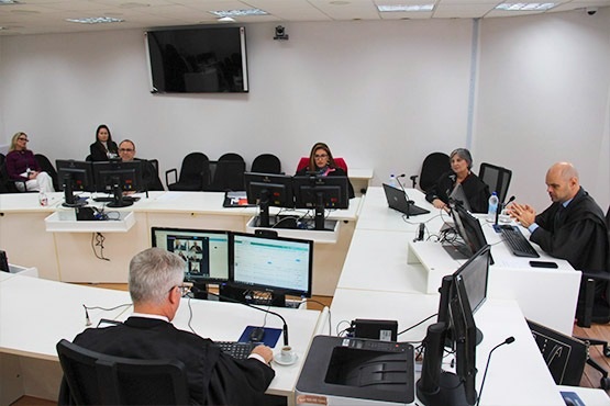 Pessoas sentadas em mesas brancas que juntas formam um retângulo. Os cinco magistrados que aparecem na foto estão espalhados pelas mesas e mexem em computadores.