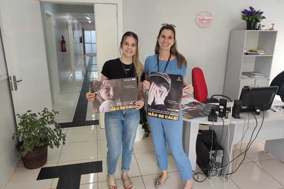 Duas mulheres posam para a foto. Cada uma segura um cartaz da campanha Maio Laranja.