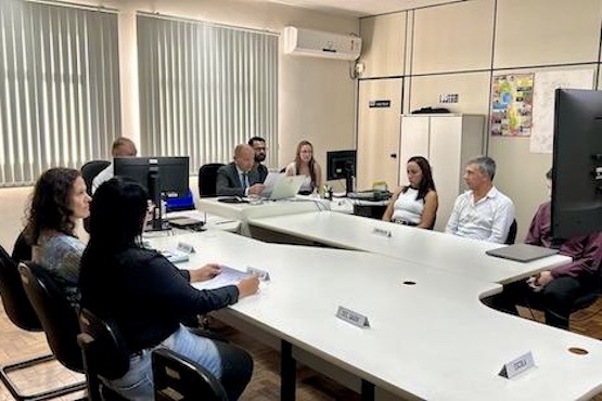 A imagem mostra uma sala de reunião com várias pessoas sentadas ao redor de uma mesa em formato de "U". Há nove pessoas ao todo, incluindo homens e mulheres. Alguns deles estão usando computadores e laptops. A sala tem paredes claras, janelas com persianas verticais fechadas e um aparelho de ar-condicionado na parede. Há também um armário branco ao fundo e um quadro de avisos na parede.
