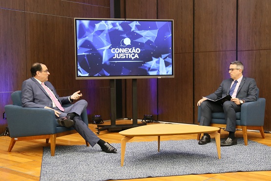 Foto tirada do programa Conexão Justiça. Dois homens sentados em poltronas baixas da cor azul escura conversam. Entre os homens está uma mesa de centro feita de madeira. Ao fundo, está uma tela com o logotipo do Conexão Justiça e algumas luzes que saem do chão na cor azul.