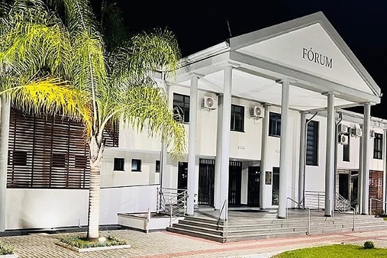 Foto lateral tirada da fachada do Fórum da comarca de Correia Pinto. A edificação de dois andares possui colunas com pé direito alto próximo da porta de entrada e palmeiras no jardim.