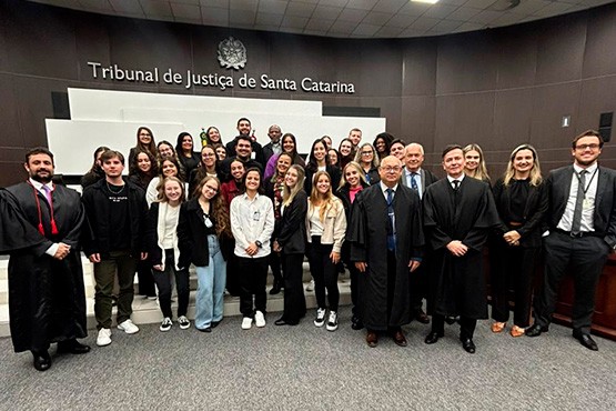 A imagem mostra um grupo de pessoas posando para uma foto no pleno do Tribunal de Justiça de Santa Catarina. Ao fundo, há uma parede com o texto "Tribunal de Justiça de Santa Catarina" e o brasão do estado. O grupo é composto por cerca de 30 pessoas, incluindo homens e mulheres de diferentes idades. Alguns estão vestidos de forma casual, enquanto outros usam trajes formais, como ternos e togas. Na frente, há alguns homens de terno e toga, sugerindo que são autoridades do tribunal.