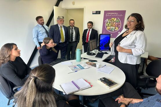 Grupo de pessoas, reunidos em uma sala, 4 pessoas em pé e 5 sentadas em cadeiras ao redor de uma mesa redonda