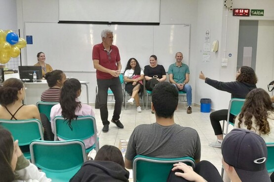 A imagem mostra uma sala de aula com várias pessoas. Na frente da sala, há um homem de cabelos grisalhos e camiseta vermelha em pé, parecendo falar ou interagir com o grupo. Atrás dele, há quatro pessoas sentadas em cadeiras, três mulheres e um homem, todos sorrindo. À esquerda, há uma mesa com um computador e balões amarelos e azuis. Os alunos estão sentados em cadeiras verdes, voltados para a frente, prestando atenção. No canto superior direito, há um relógio digital e uma placa de "Saída".