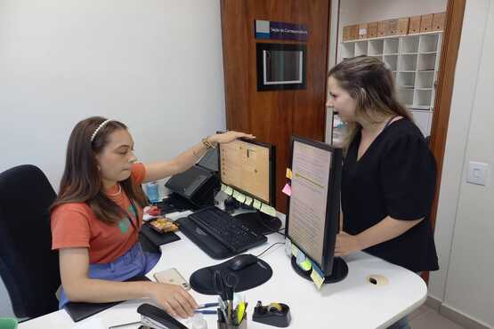 Foto de duas mulheres. A mulher localizada mais à esquerda da fotografia e que usa uma camiseta laranja está sentada em uma mesa branca e mexe em um dos dois monitores a sua frente. A mulher à direita está de camiseta preta e acompanha a movimentação.