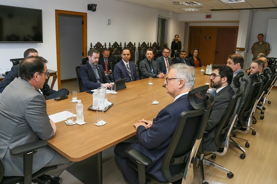 A imagem mostra uma reunião formal em uma sala de conferências. Há uma mesa retangular de madeira clara, ao redor da qual estão sentadas 14 pessoas, todas vestidas formalmente com ternos e gravatas. A maioria dos participantes são homens, com apenas uma mulher visível ao fundo. Na mesa, há garrafas de água, copos e alguns papéis. A sala tem paredes brancas, uma televisão de tela plana na parede à esquerda e uma porta de madeira ao fundo. Algumas cadeiras estofadas com encosto alto estão dispostas ao redor da mesa.