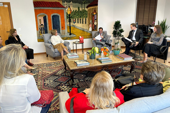 Foto tirada de homens e mulheres com roupas sociais sentados em sofás e poltronas ao redor de uma mesa de centro, que possui livros e flores em cima.