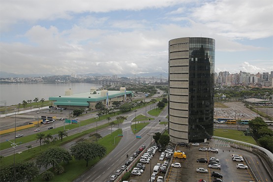 Foto aérea de prédio redondo e espelhado. Ao lado estão ruas. Em frente há um estacionamento com carros parados.