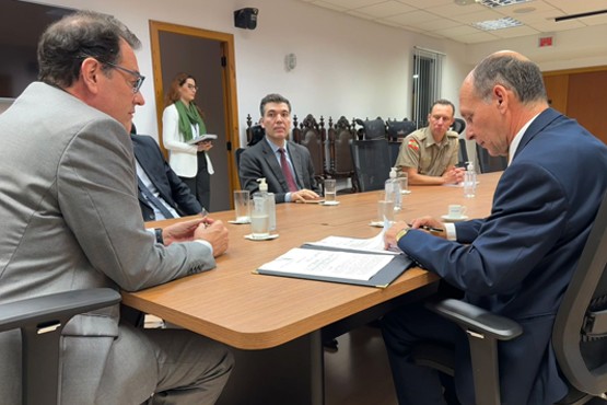 Foto de 5 homens, sendo 4 de terno e gravata, e um com a farda da Polícia Militar, dentro de uma sala, ao redor de uma mesa retangular e sobre ela estão alguns copos de água, xícaras de café e algumas embalagens de álcool em gel. Ao fundo está uma mulher em pé, passando pela entrada de uma porta.