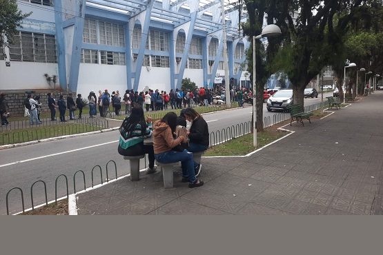 A imagem mostra 3 meninas sentadas em um banco de cimento, na calçada em frente ao complexo. O fundo mostra a rua e os carros estacionados, a fachada do completo e algumas árvores.