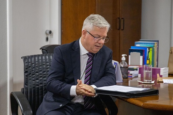 Foto da parte de uma mesa em que está um homem, de terno e gravata, sentado em uma cadeira, lendo um papel. Sobre a mesa, também está um copo de água e alguns pertences pessoais.