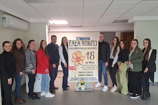 A imagem mostra um grupo de 11 pessoas posando para uma foto em um ambiente interno. Eles estão em pé, lado a lado, em frente a um cartaz que diz "Faça Bonito - Proteja nossas crianças e adolescentes". O cartaz também menciona a data "18 de Maio" e destaca a importância de "Esquecer é Permitir, Lembrar é Combater". O grupo é composto por homens e mulheres de diferentes idades, todos sorrindo para a câmera. No chão, em frente ao cartaz, há um pequeno arranjo de flores.