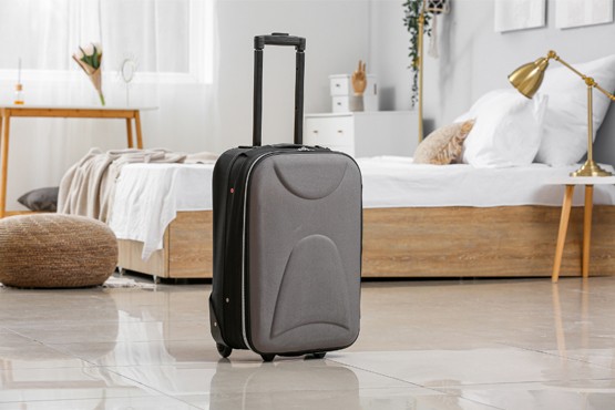 Imagem de uma mala de viagem cinza com a alça levantada. Ao fundo alguns móveis como uma cama e uma mesinha de lateral.