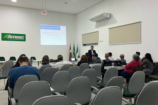 Foto de um grupo de pessoas em uma sala, sentadas em cadeiras dispostas de forma enfileiradas. Em frente está em uma parede, a projeção de um slide com o título “Grupos reflexivos”. No chão, estão alguns mastros com algumas bandeiras.