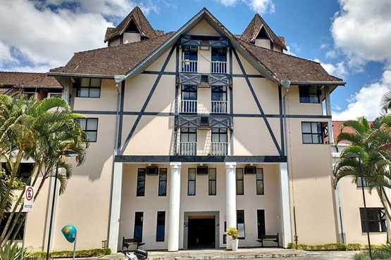 Fórum Central do TJ da comarca de Blumenau. A construção é estilo chaimel com quatro pilares na parte da entrada do local. A imagem também mostra parte do céu com nuvens e algumas palmeiras junto ao gramado verde.