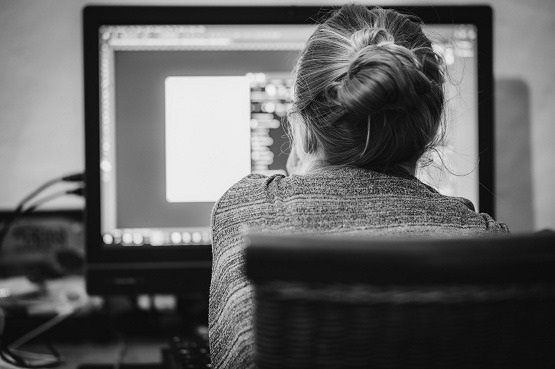 Uma mulher está sentada em uma cadeira de costas para a foto, ela está virada e mexendo em um computador. A foto está em preto e branco.