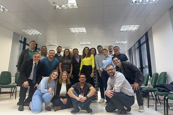 Grupo de pessoas estão em uma sala, e posam para uma foto em grupo. Algumas estão em pé, outras estão agachadas ou sentadas no chão.