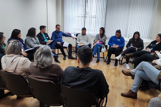 Um grupo de pessoas estão em uma sala sentados em cadeiras organizadas em um círculo, todos estão com caderno e caneta nas mãos