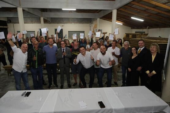 Grupo de pessoas estão em uma sala e posam para uma foto em grupo. Todos estão em pé e enfileirados. Alguns deles estão levantando uma folha de papel na mão.