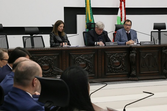 A imagem mostra uma reunião. Três pessoas estão sentadas atrás de uma mesa de madeira esculpida, que possui detalhes ornamentais. A pessoa no centro é um homem de cabelos grisalhos, usando óculos e um terno escuro. À sua esquerda, há uma mulher de cabelos castanhos, também vestindo roupas formais. À direita, outro homem de óculos e terno azul. Atrás deles, há bandeiras do Brasil e de Santa Catarina. Na frente da mesa, outras pessoas estão sentadas, mas suas faces não são claramente visíveis.