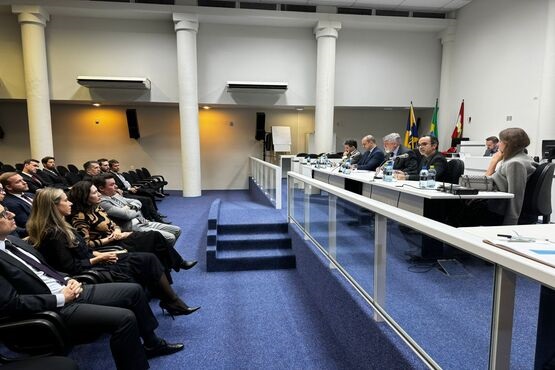 Em uma sala, um grupo de pessoas estão sentados em cadeiras em fileiras, de frente com eles estã outras pessoas, também sentadas em cadeiras, e com uma mesa entre eles.
