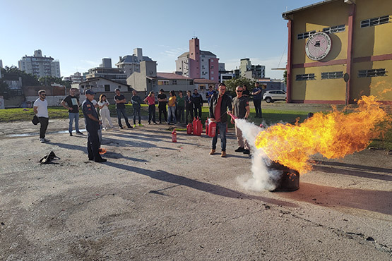 Foto tirada de um homem utilizando um extintor vermelho para apagar o fogo dentro de um tonel. Tanto à direita quanto à esquerda do homem estão outras duas figuras masculinas para auxiliar. O homem à esquerda é um bombeiro fardado. Ao fundo da imagem, pessoas observam a ação.