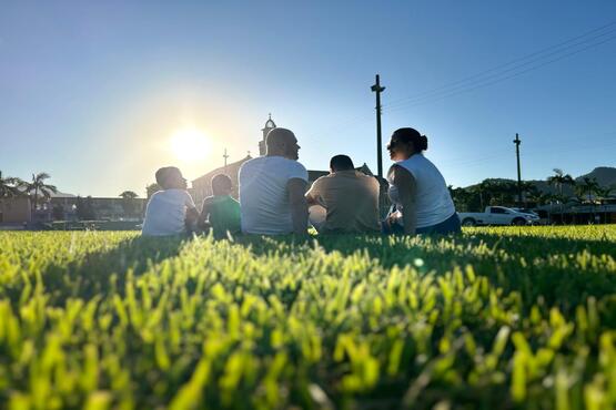 Uma família composta por cinco pessoas está sentada em um gramado de costas para a fotografia.