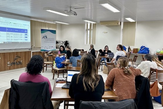 Pessoas sentadas em mesas estão de costas para a foto e acompanham a reunião do Programa Novos Caminhos. No fundo do lado esquerdo da fotografia está uma projeção e um banner do programa.