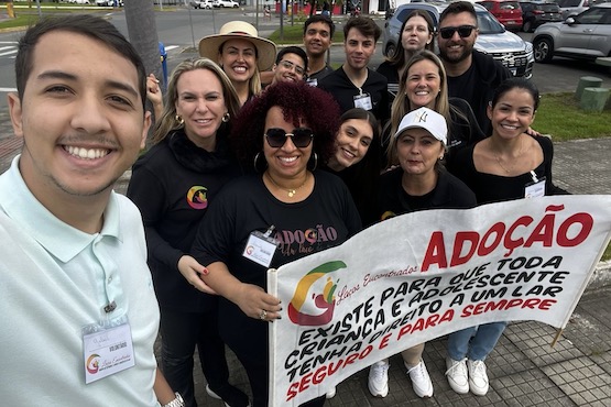 Selfie dos participantes do Grupo de Estudos e Apoio à Adoção (GEAAI). No primeiro plano e à esquerda, um homem de camiseta verde clara tira a foto. Pessoas de blusa preta, que estão localizadas mais ao fundo à direita, posam para a foto e seguram um cartaz com uma frase sobre a adoção.