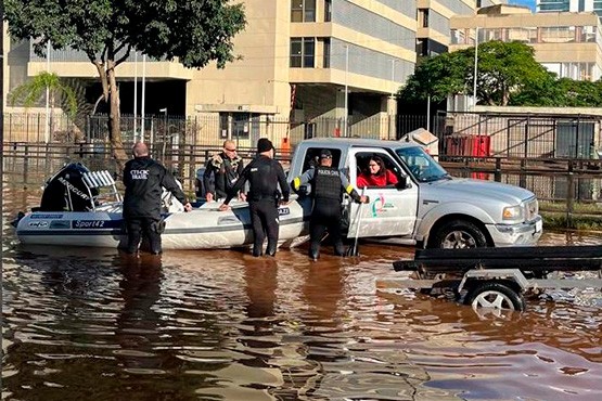 Camioneta puxa um bote em meio à enchente. Ao redor do bote tem pessoas tanto que estão sendo resgatadas quando realizando o resgate.