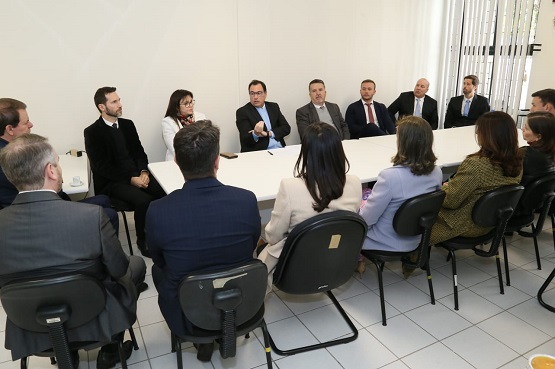 A imagem mostra uma reunião formal com 15 pessoas sentadas ao redor de uma mesa retangular branca. A maioria das pessoas está vestida com trajes formais, como ternos e blazers. A mesa está em uma sala com paredes brancas e piso de azulejo claro. As pessoas estão atentas, algumas conversando entre si, e parece que um homem no centro da mesa está falando, gesticulando com a mão direita. Ao fundo, há uma janela com persianas verticais.