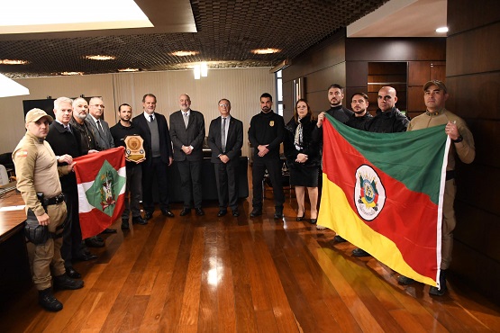 Imagem de um grupo de pessoas dentro de uma sala sendo 13 homens e uma mulher. Todos estão em pé e posam para uma foto. Quatro dos homens, seguram uma bandeira do Rio Grande do Sul e outros quatro, seguram uma bandeira de Santa Catarina.