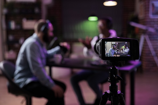 Imagem de uma câmera fotográfica, filmando duas pessoas sentadas sobre cadeiras e uma mesa redonda entre eles.