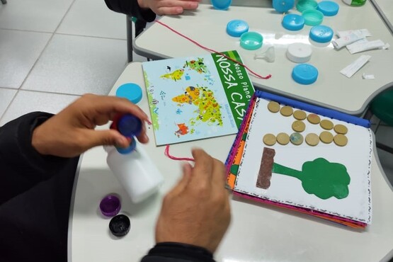 A imagem mostra uma pessoa manipulando materiais de artesanato em uma mesa. A pessoa está segurando um frasco branco com uma tampa azul. Na mesa, há vários objetos, incluindo tampas de garrafa de plástico de várias cores, um livro ou caderno com a capa escrita "Nossa Casa" e uma página com uma árvore feita de materiais colados, como tampas e recortes de papel. A mesa está em um ambiente interno, possivelmente uma sala de aula ou um espaço de atividades.