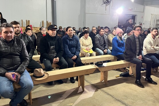Grupo de pessoas estão dentro de uma sala, sentadas em bancos compridos e enfileirados. Todas estão bem agasalhadas, com casacos e tocas.