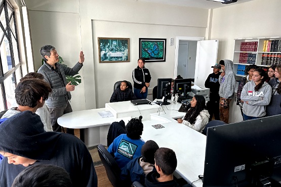 A imagem mostra uma sala com várias pessoas, a maioria jovens, prestando atenção em um homem mais velho que está em pé, falando e gesticulando com a mão levantada. Ele está à esquerda da imagem, vestindo uma jaqueta cinza. A sala tem paredes brancas e duas janelas grandes à esquerda, que deixam entrar bastante luz natural. Há duas mesas brancas no centro, com alguns computadores e papéis. Algumas pessoas estão sentadas ao redor das mesas, enquanto outras estão em pé ao fundo e à direita. Na parede ao fundo, há duas pinturas emolduradas e uma estante com livros à direita.