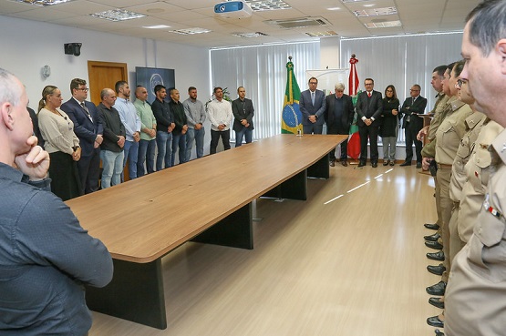 A imagem mostra uma sala de reuniões com uma grande mesa de madeira no centro. Ao redor da mesa, há várias pessoas em pé, divididas em dois grupos. À esquerda, há um grupo de homens e mulheres vestidos de maneira casual e formal. À direita, há um grupo de homens e mulheres fardados. No fundo da sala, há duas bandeiras, uma do Brasil e outra de Santa Catarina, além de um banner do PJSC. A sala tem piso de madeira clara e janelas com persianas brancas.