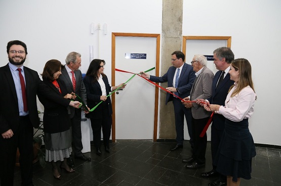 A imagem mostra um grupo de oito pessoas participando de uma cerimônia de inauguração. Eles estão em frente a uma porta branca com uma placa azul, e estão cortando uma fita vermelha e verde. As pessoas estão vestidas formalmente, com ternos e roupas sociais. Há um homem de barba e óculos à esquerda, sorrindo, e os outros estão concentrados na fita. A parede ao fundo é branca, e há uma coluna de concreto entre duas portas.