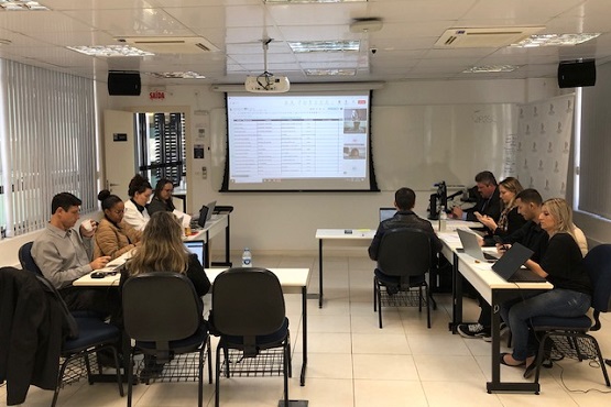 A imagem mostra uma sala de reunião com várias pessoas sentadas em torno de duas mesas retangulares dispostas em formato de "U". Há um total de 10 pessoas, todas concentradas em laptops ou documentos. Na parede ao fundo, há uma tela de projeção mostrando uma planilha com várias colunas e linhas. A sala é bem iluminada, com luz natural entrando pelas janelas à esquerda, que têm persianas verticais. O teto é branco com luminárias embutidas.