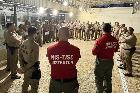 Homens fardados parados em círculo. No primeiro plano estão dois homens de costas para a foto com blusas vermelhas escrito em letras brancas “NIS-TJSC INSTRUTOR”. O restante dos homens usam fardas bege.