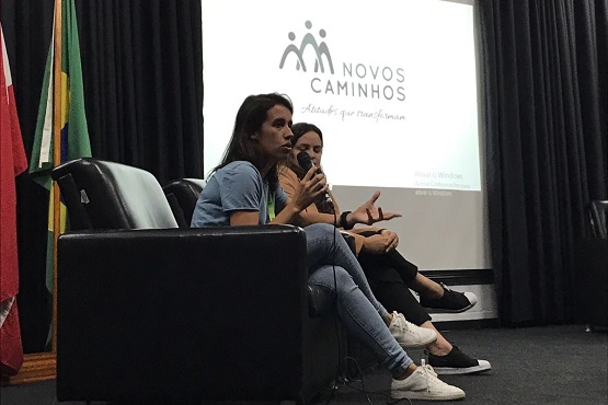 A imagem mostra duas mulheres sentadas em poltronas pretas em um palco, participando de um evento do Programa Novos Caminhos. A mulher à esquerda está falando ao microfone e usa uma camiseta azul clara e calça jeans. A mulher à direita está prestando atenção na fala. Atrás delas, há uma tela de projeção com o logotipo "Novos Caminhos" e o slogan "Atitudes que transformam". Ao lado da tela, há bandeiras, incluindo a bandeira do Brasil.