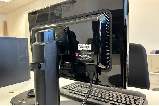A imagem mostra a parte traseira de um monitor LG. O monitor está preso a um suporte articulado, que parece ser ajustável. Há cabos conectados na parte de trás do monitor, incluindo um cabo HDMI e um cabo de energia. Abaixo do monitor, é possível ver parcialmente um teclado.