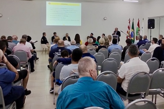 A imagem mostra uma sala de conferências com várias pessoas sentadas em cadeiras, todas voltadas para a frente da sala. Na frente, há algumas pessoas em pé, possivelmente palestrantes ou organizadores, e uma tela de projeção com algo exibido, mas o conteúdo não está legível na foto. Há também bandeiras ao lado direito da sala, incluindo a bandeira do Brasil. A sala parece estar bem iluminada e as pessoas estão prestando atenção ao que está sendo apresentado.