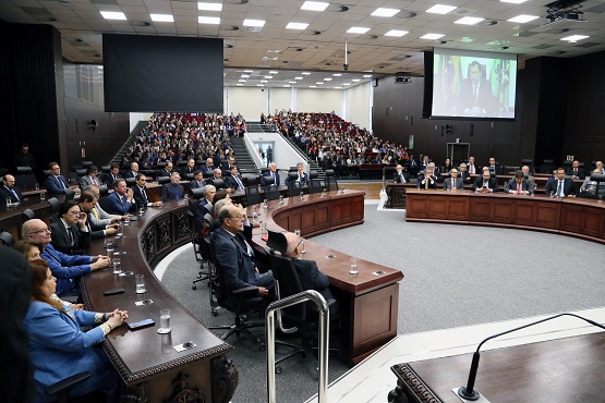 A imagem mostra uma sessão em um auditório ou plenário, possivelmente de um órgão legislativo ou uma conferência. Há várias pessoas sentadas em mesas dispostas em formato de "U" ou semicírculo, voltadas para um ponto central. As pessoas estão vestidas formalmente, muitas usando ternos. Ao fundo, há uma plateia cheia de pessoas sentadas, e na parede de trás, há uma grande tela de projeção. O ambiente é bem iluminado e parece ser um evento formal ou uma reunião importante.