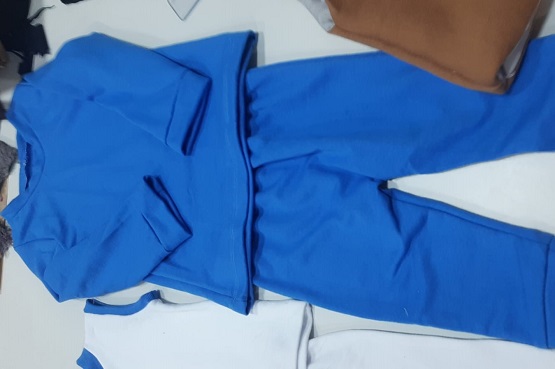 A imagem mostra um conjunto de roupas infantis de cor azul. O conjunto é composto por uma blusa de manga longa e uma calça. Ao lado do conjunto azul, há outras peças de roupa, incluindo uma peça branca com detalhes azuis e outra peça de cor marrom. As roupas estão dispostas sobre uma superfície clara.
